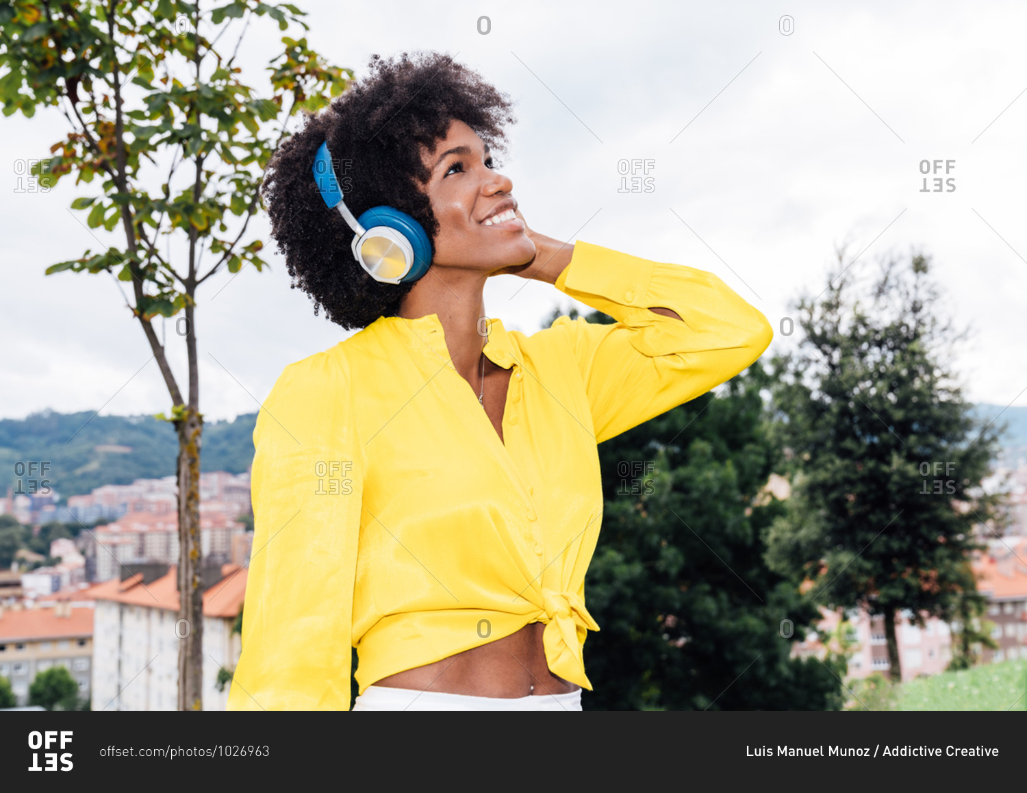 Cheerful black female smiling and listening to music in headphones while resting in park in summer