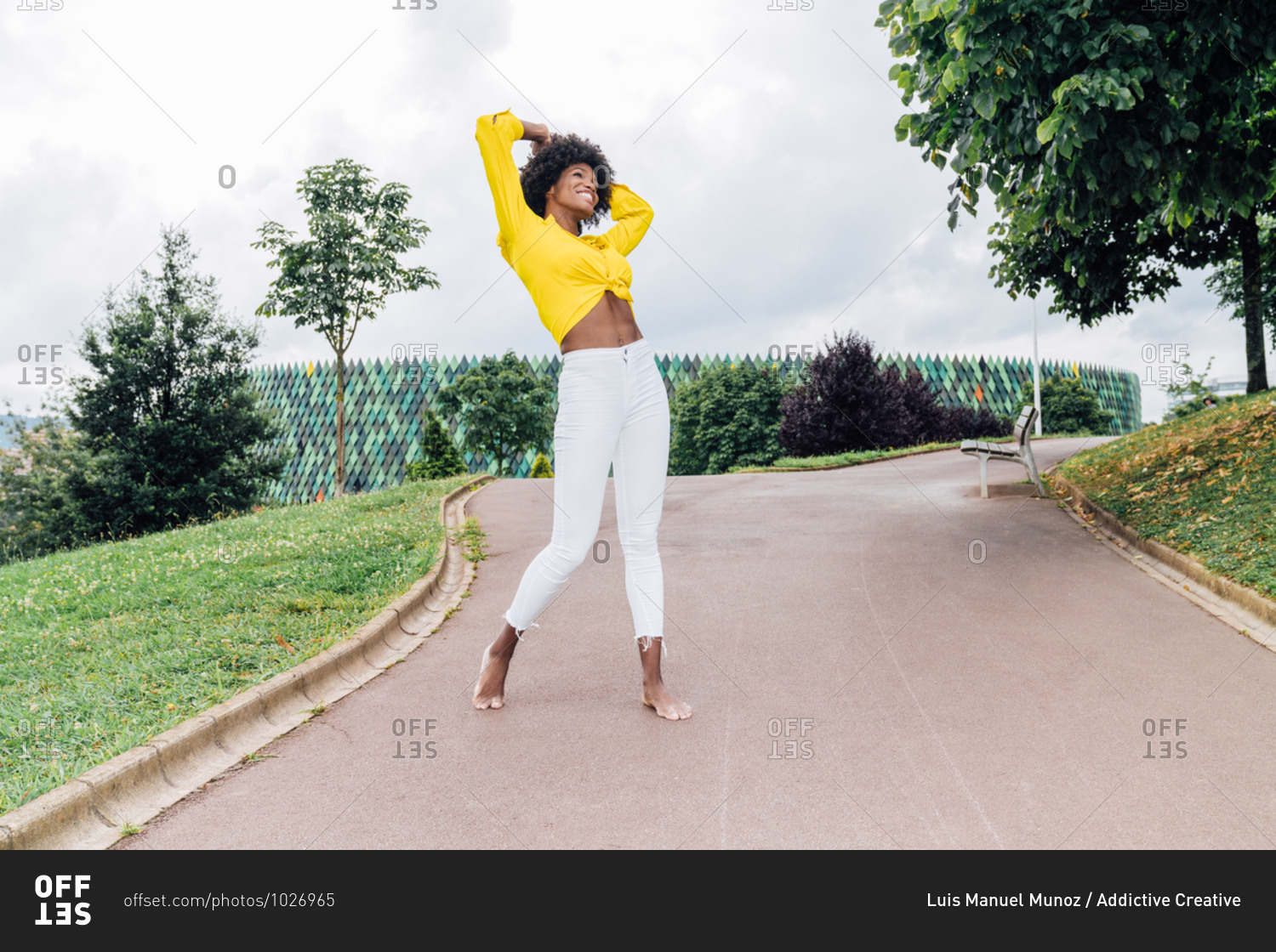 From below optimistic black woman in trendy clothes smiling and dancing with raised arms on sunny day in park