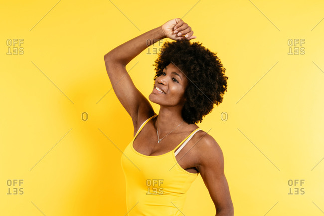 Positive African American female with Afro smiling and raising arm while standing against yellow background
