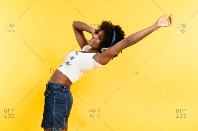 Glad African American female smiling and listening to music in headphones with closed eyes against yellow background