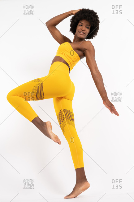 Low angle of fit black female in yellow sportswear balancing on one leg and looking away against gray background