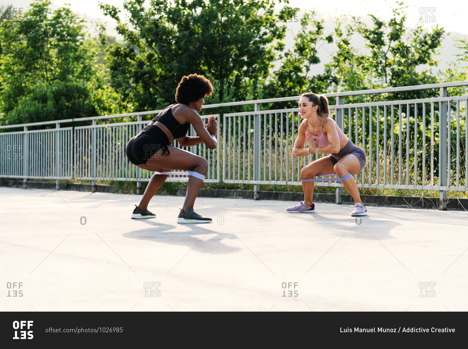 Full body of determined young multiethnic sportswomen doing squat exercise with resistance band during fitness workout in park in summer day