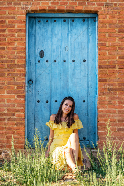 Charming female in trendy dress sitting on shabby wooden doors of building while looking at camera and relaxing during weekend in city
