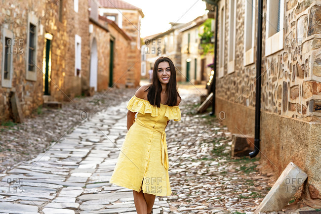 Carefree female in summer dress standing on narrow paved street of historic town and looking at camera