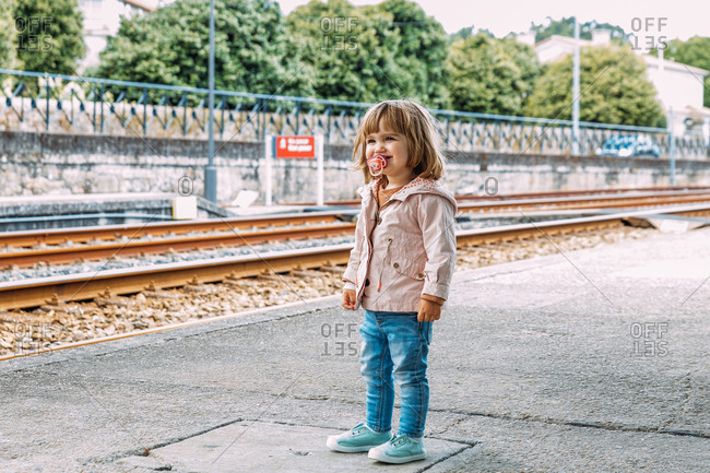 Positive adorable little girl in casual wear standing at railway station and looking away while waiting for train