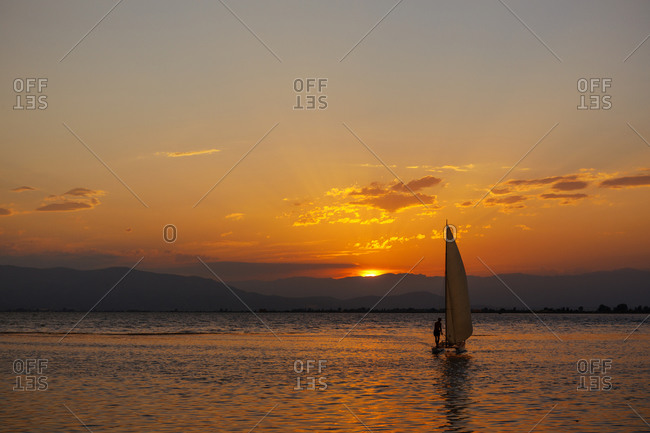 Little sailing boat in the sea at sunset with the sun and clouds in the background. Sailing on a summer adventure in the Mediterranean sea
