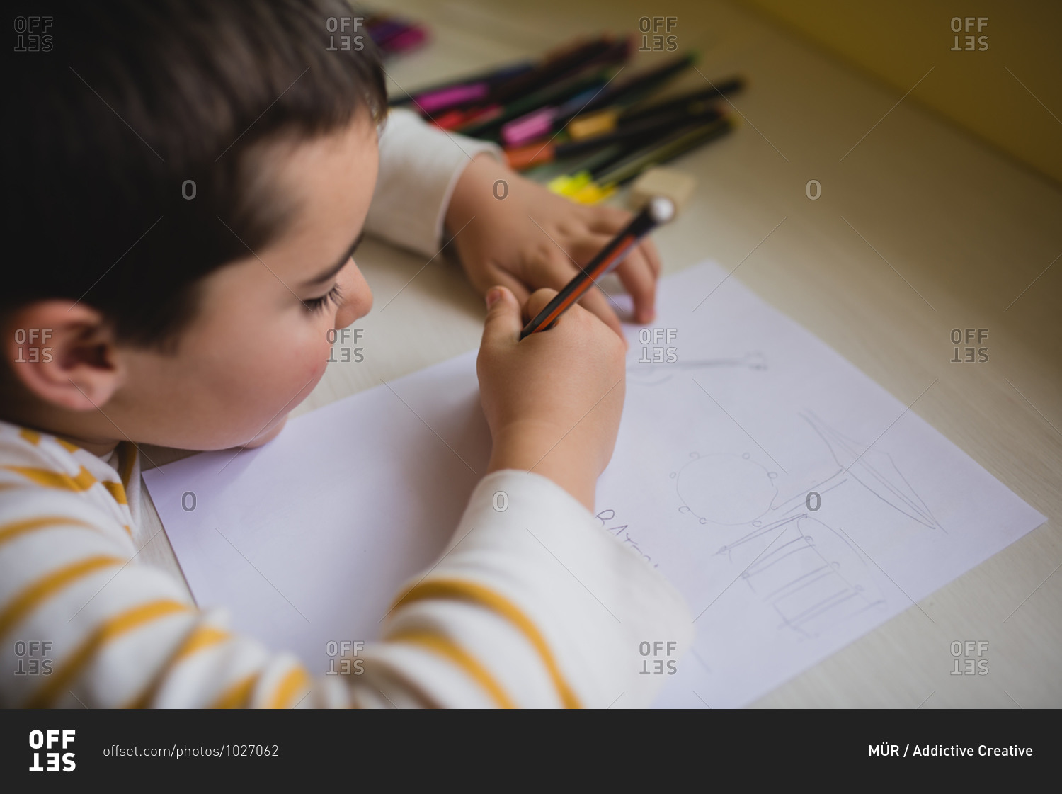 Side view of crop cute kid sitting at wooden table and drawing with pencil on paper while relaxing at weekend at home