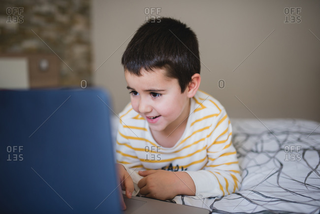 Focused little boy in casual wear browsing laptop while spending free time in bedroom at home