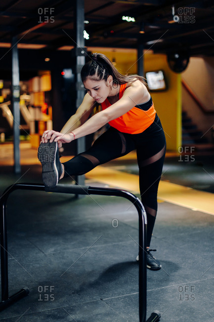 Full body young sporty female in fitness outfit performing stretching exercise near metal bar while warming up during training in gym