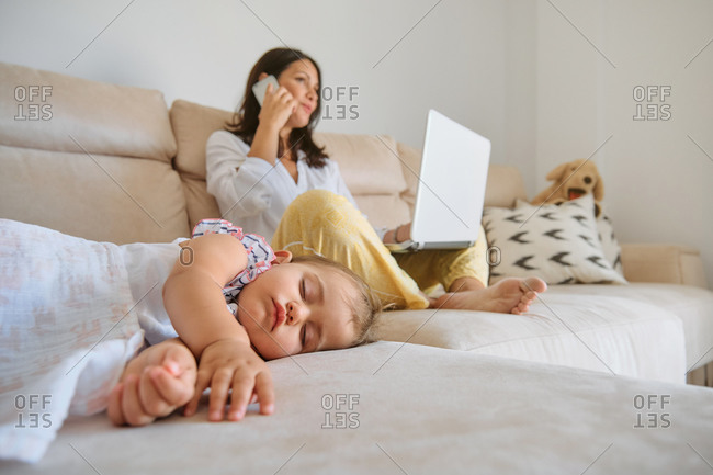 Little girl sleeping on the sofa while her mother works with a laptop and talking with the mobile phone next to her