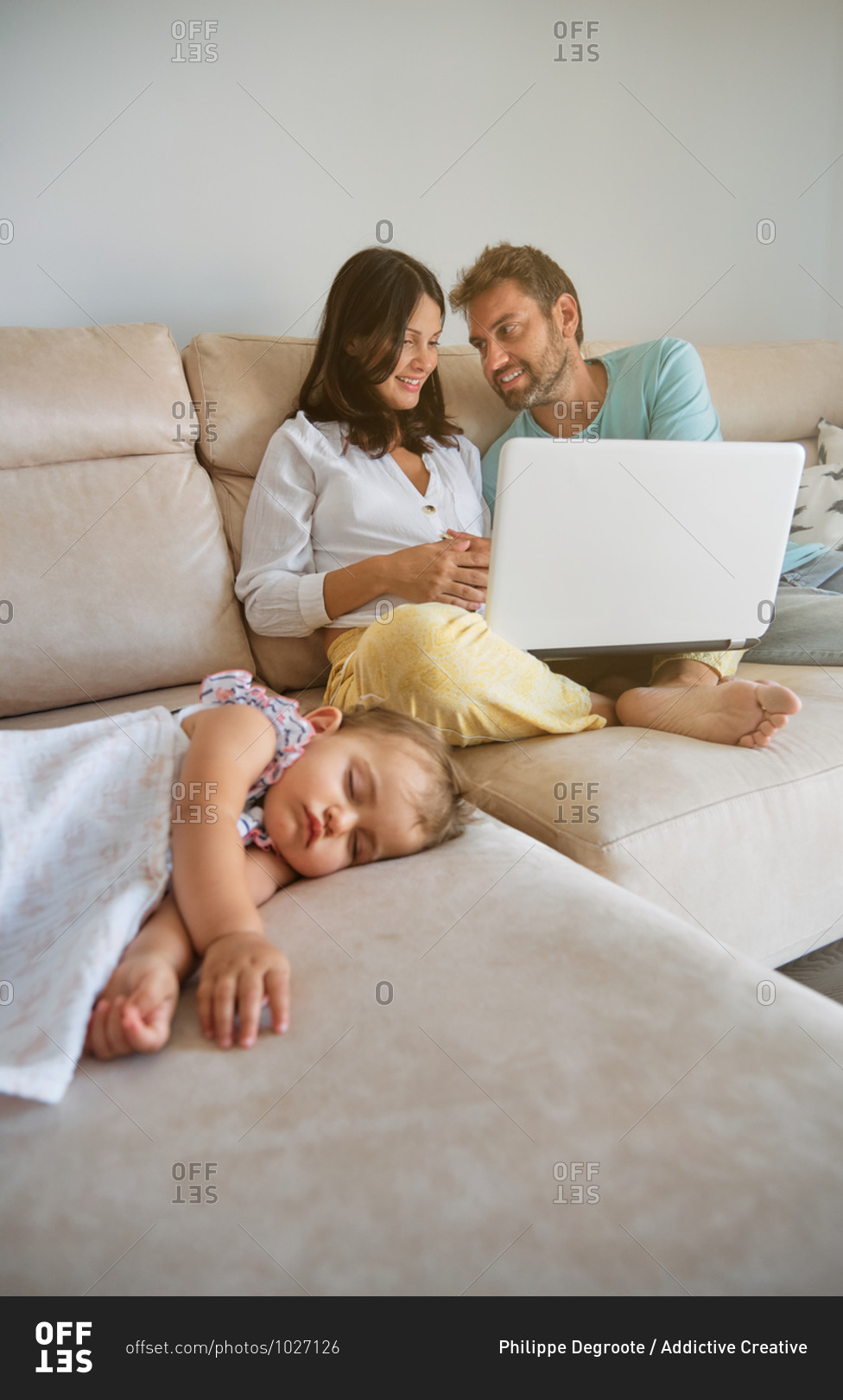 Little girl sleeping on the couch while a pregnant couple is distracted with a laptop next to her at home