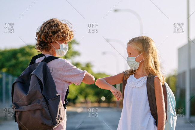Two kids with school bags and masks greeting each other with a gesture of touching elbows in the street in front of a park