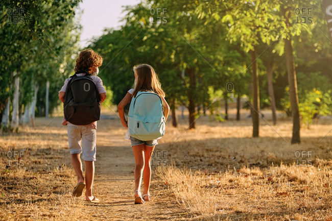 Two kids with school bags and a mask on their backs walking along a path in the woods while talking