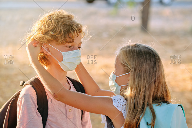 Blond little girl with a school bag and a mask helping a boy to put on her mask outdoors