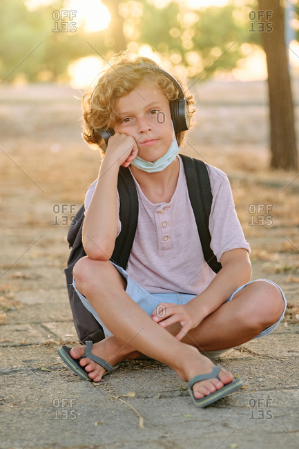 Vertical photo of a blond boy with the mask down and school bag sitting while listening to music with his hand resting on his chin and bored expression facing the camera