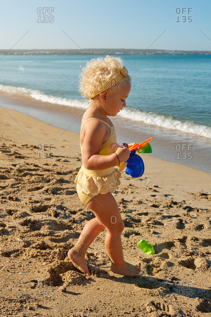 Vertical photo of a little blonde girl in a yellow striped swimsuit walking on the beach with plastic toys in her hand