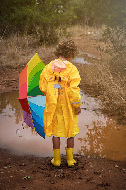 Vertical photo of a blond boy on his back in a yellow raincoat with a hood with a donkey and yellow water boots and a colorful umbrella in the rain in a puddle in the forest
