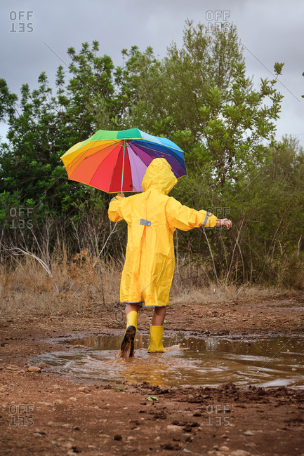 Vertical photo of a child on his back with a yellow raincoat and a colorful umbrella playing in a puddle with rain boots in the middle of the forest