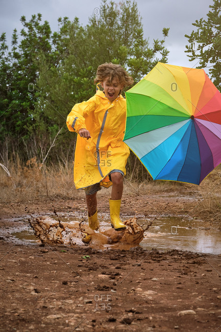 Vertical photo of a child with a yellow raincoat and a colorful umbrella playing in a puddle splashing the water with the rain boots in the middle of the forest
