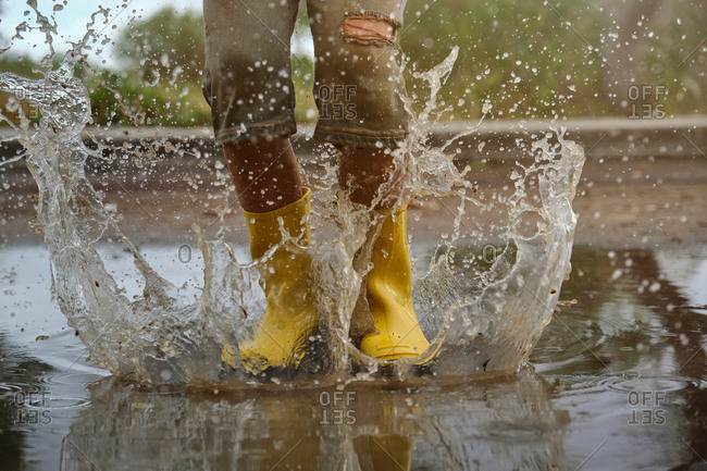 Detail of yellow rain boots hitting a puddle splashing water in a forest