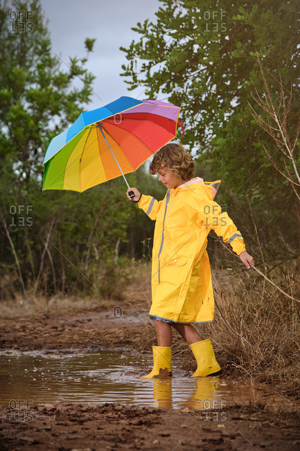 Vertical photo of a blond boy with a yellow raincoat, rain boots and a colorful umbrella playing with a stick and a puddle in the forest