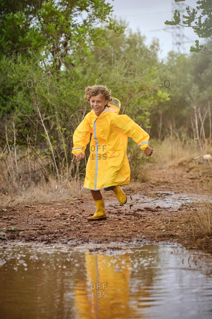 Vertical photo of a blond boy in a raincoat and yellow rain boots running along a path full of puddles in the forest while smiling