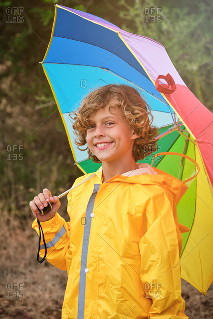 Vertical photo of a blond boy with curly hair in a yellow raincoat holding a colored umbrella while smiling on camera in the middle of a forest