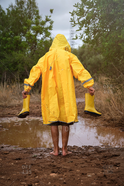 Vertical photo of a child in her back with yellow raincoat walking on a barefoot muddy path with water boots in his hands in a forest