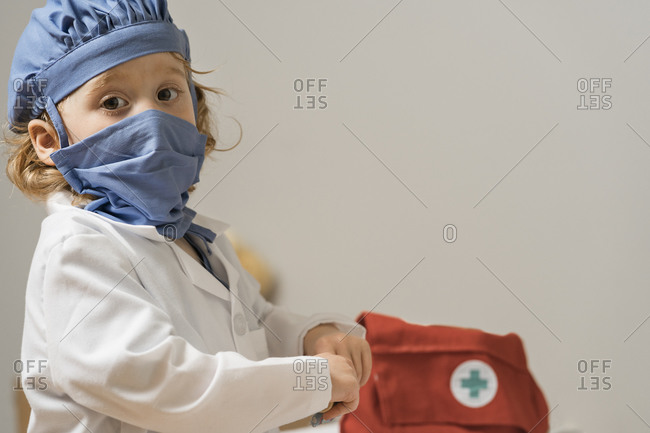 Young child wearing medical PPE turns to look directly at viewer as medical bag sits in the background