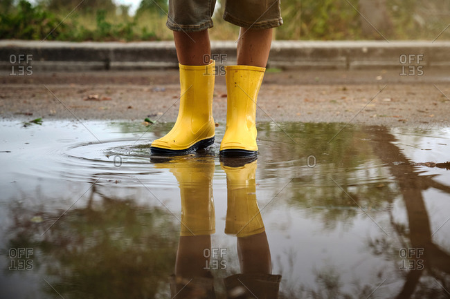 Detail of the legs of a boy with yellow rain boots in a puddle with the reflection in the water in a path in the forest
