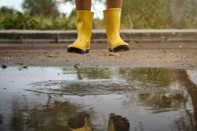 Detail of the legs of a boy with yellow rain boots jumping above a puddle water in a path in the forest
