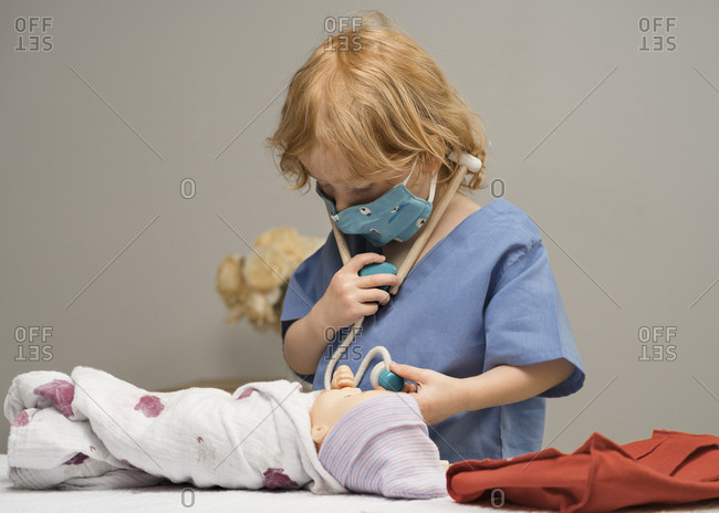 Young child wearing medical PPE examines a baby doll by checking its vitals with a stethoscope