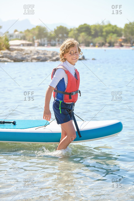 Vertical photo of a blond teenager with curly hair and life vest dragging a paddleboard across the water of the sea while smiling at the camera