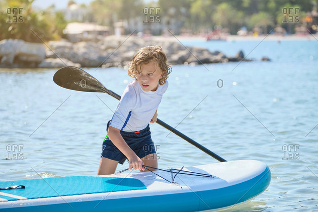Blond teenager with curly hair dragging a paddleboard across the water of the sea in a sunny day
