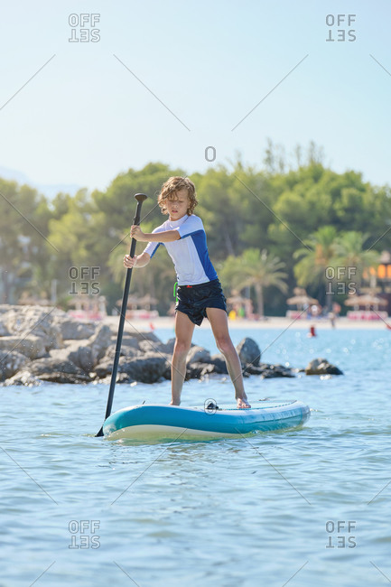 Vertical photo of a child standing on a paddle surfboard rowing in the middle of the sea water with the beach on the background