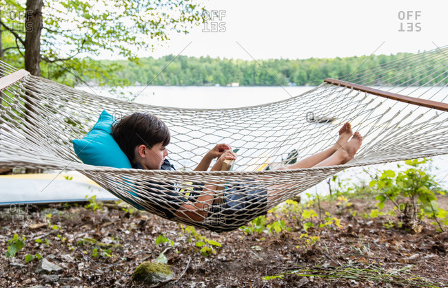 Young boy laying in a woven hammock relaxing and reading a book.