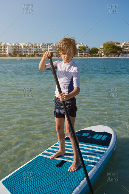 Vertical photo of a boy standing on a paddle surfboard rowing in the middle of the sea water with the beach with buildings on the background