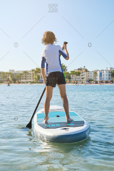 Vertical photo of a blonde boy in his back on a paddle surfboard rowing in the middle of the sea with the beach on the background