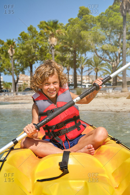 Vertical photo of a blond boy with curly hair and a life jacket rowing and facing the camera in a yellow kayak in the sea with the beach in the background on a sunny day
