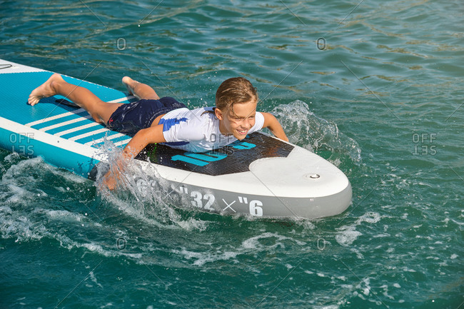 Blond boy lying on a paddle board rowing with his hands splashing the waves in a sunny day