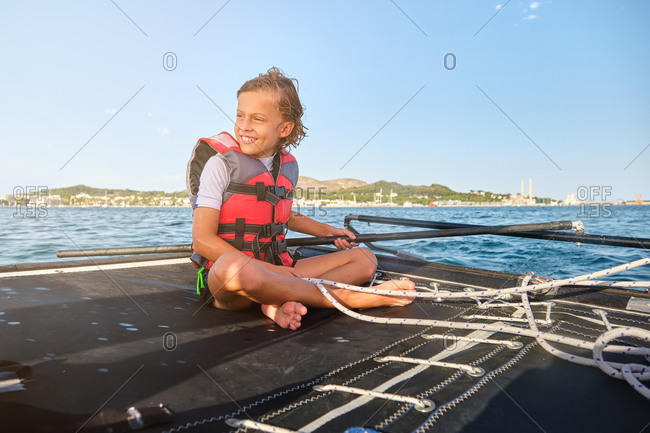 Blond kid with life jacket sitting in a boat holding the rudder and a rope to drive the boat while smiling with the shore far away in the background