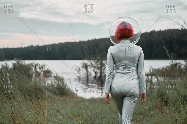Back view of unrecognizable futuristic young red haired female in silver space suit and glass helmet walking to flooding river
