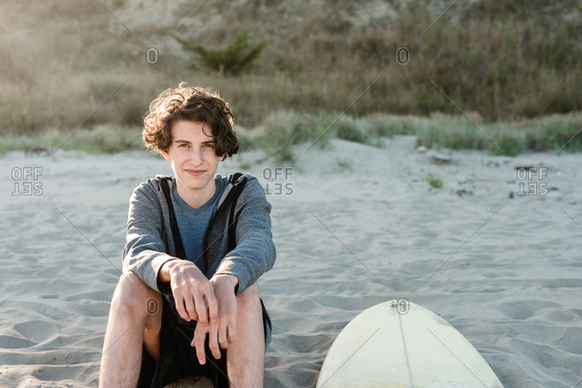 Long-haired teenager sitting next to surfboard on a beach in New Zealand