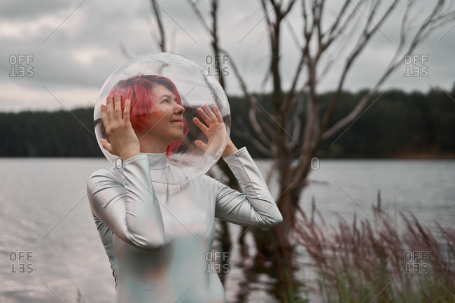 Futuristic young red haired female in silver space suit and glass helmet walking near flooding river