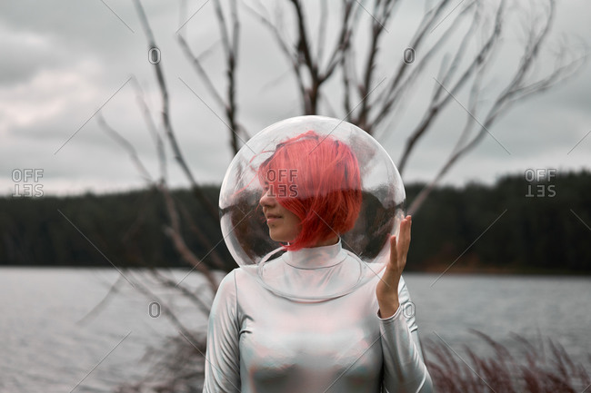 Futuristic young red haired female in silver space suit and glass helmet walking near flooding river