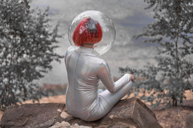 Back view of unrecognizable futuristic young red haired female in silver space suit and glass helmet sitting near flooding river