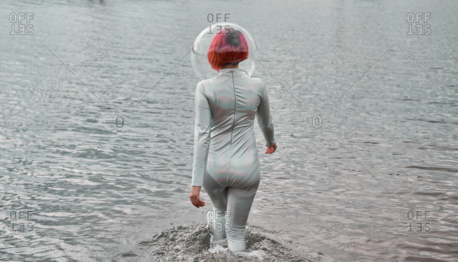 Back view of unrecognizable futuristic young red haired female in silver space suit and glass helmet walking on flooding river