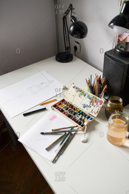 High angle of various paintbrushes and watercolor palette arranged on table with paper and glass jar of water in art studio
