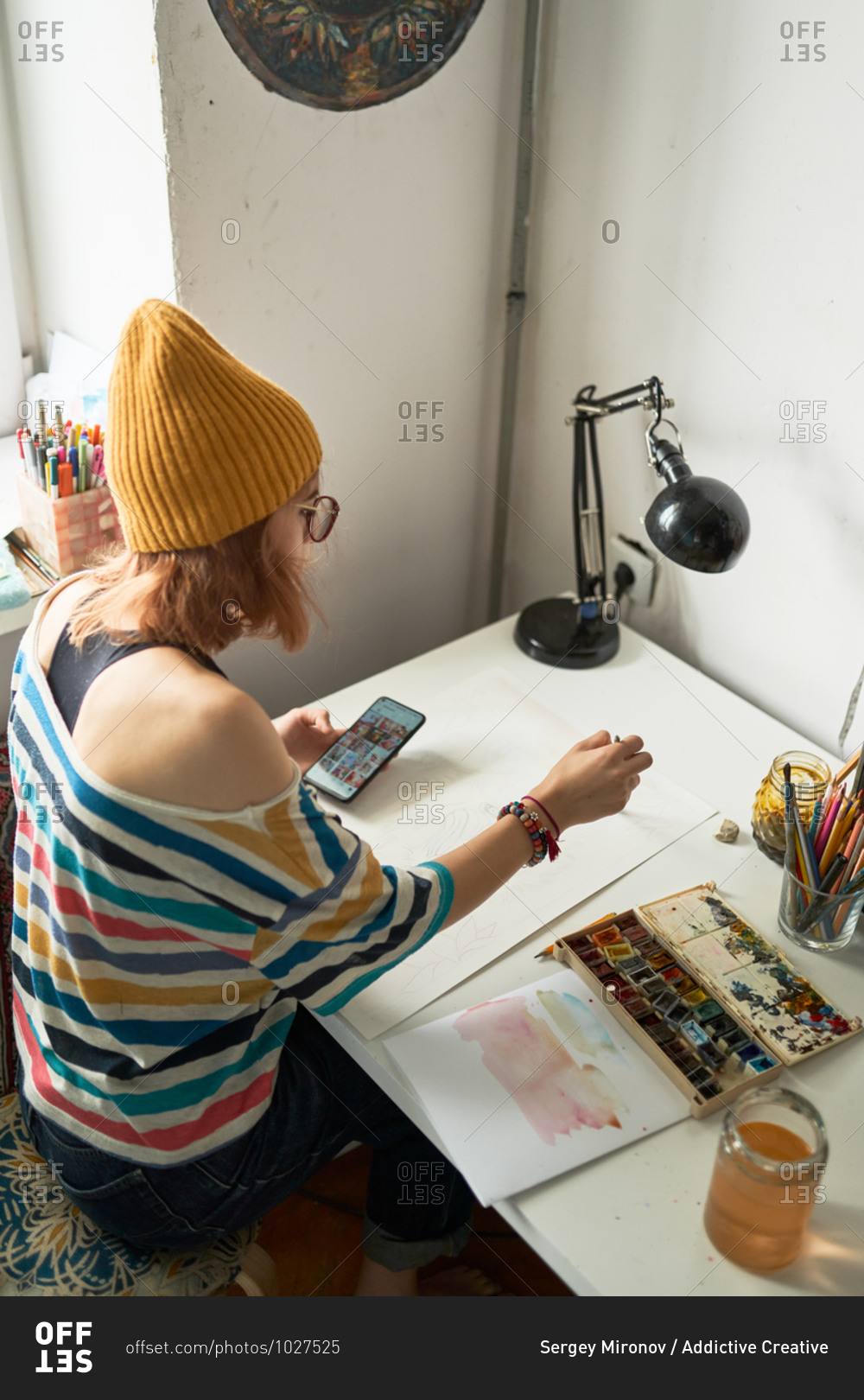 Female artist painting with watercolors and watching online video tutorial on smartphone while creating artwork in studio