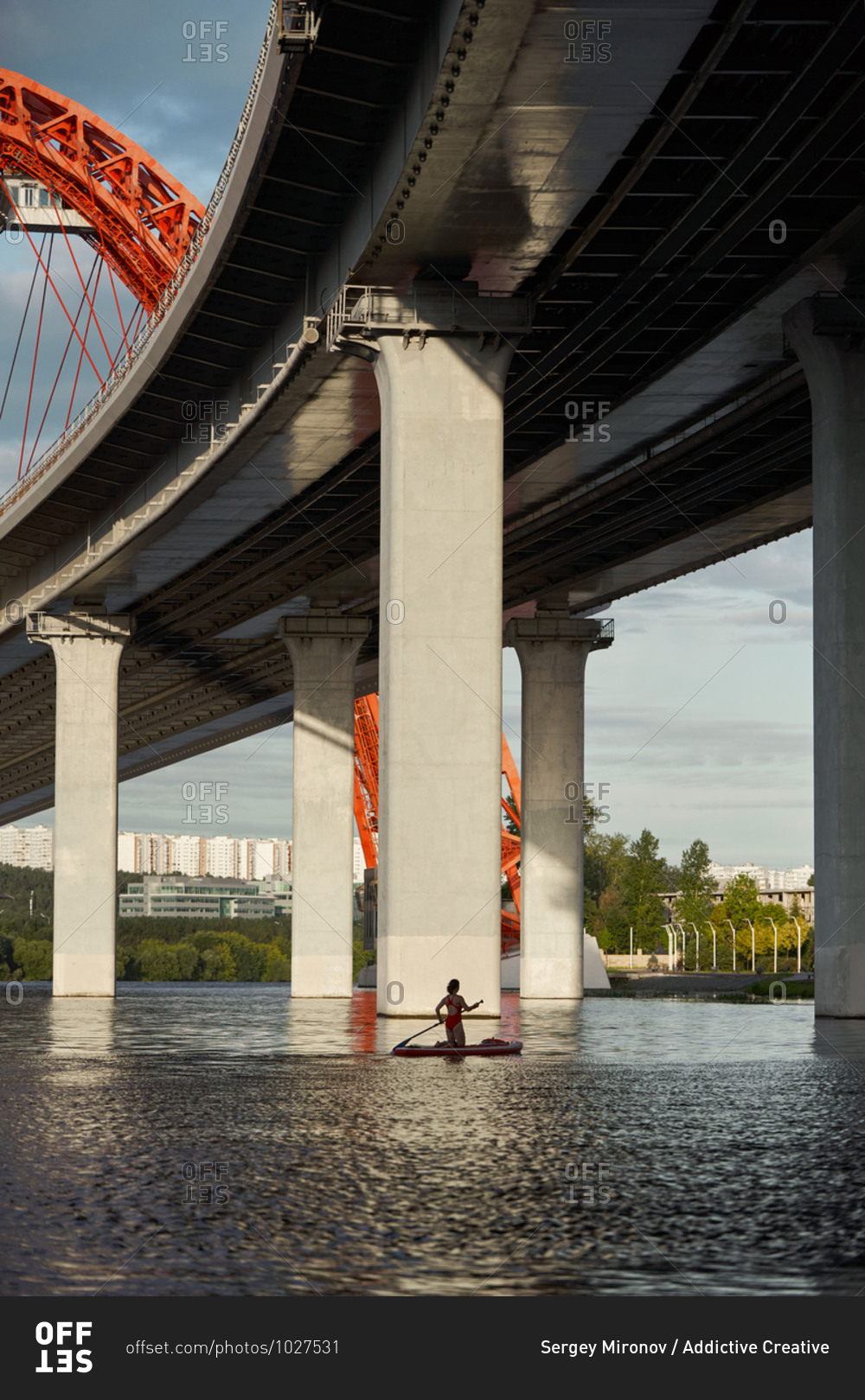 Side view of woman standing on paddle board in river and rowing with paddle under bridge in city